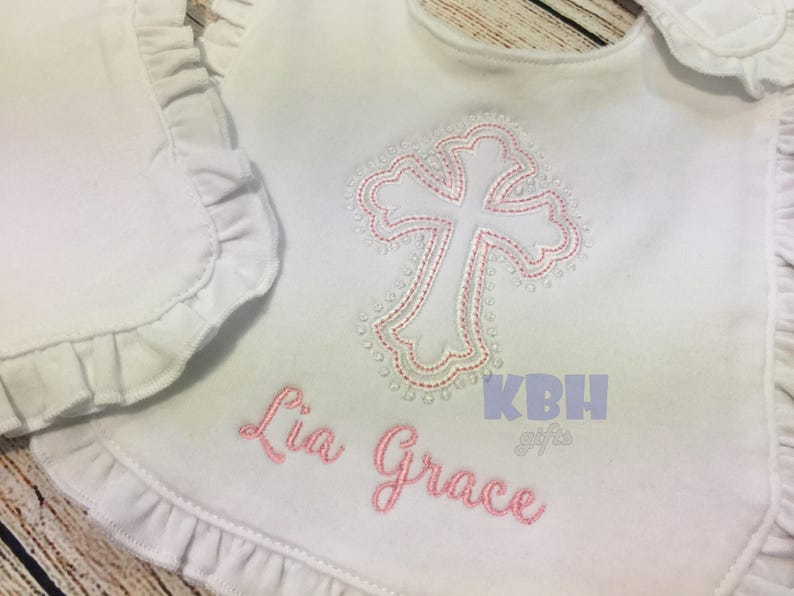 Puede incluir: Babero blanco con una cruz bordada en rosa y el nombre "Lia Grace" sobre un fondo blanco.
