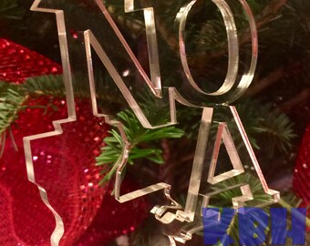 May include: Clear acrylic Christmas ornament shaped like the state of Louisiana with the letters "NO" and "LA" inside. The ornament is hanging on a Christmas tree with red ornaments.