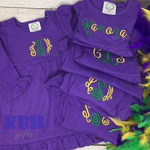 May include: A set of five purple long-sleeved dresses with green and yellow embroidered monograms. The dresses are layered on top of each other. The top dress has the name "Ramona" embroidered on it. The bottom dress has the text "KBH gifts" embroidered on it.