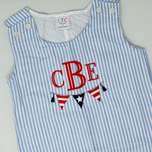 Puede incluir: Mono azul y blanco a rayas con las letras "CBE" bordadas en rojo y una guirnalda roja, blanca y azul con borlas.