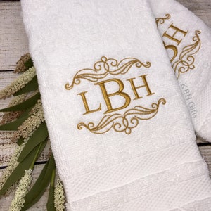May include: Two white bath towels with gold embroidered monograms. The monogram on the left towel is "LBH" and the monogram on the right towel is "DH".