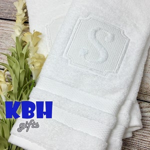 May include: Two white bath towels with a monogrammed letter "S" embroidered in the center of the top towel. The towels are folded and stacked on a wooden surface.