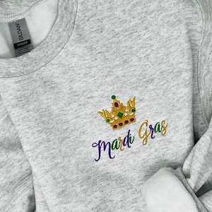 May include: A grey sweatshirt with an embroidered design of a gold crown with purple and green accents and the text "Mardi Gras" in purple.