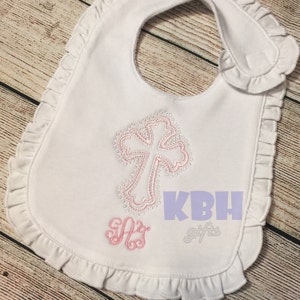 Puede incluir: Babero blanco con ribete de volantes rosa y una cruz bordada en rosa con un contorno de perlas. El babero tambi&eacute;n tiene un monograma bordado en rosa "SAS" y el texto "KBH gifts" en morado.