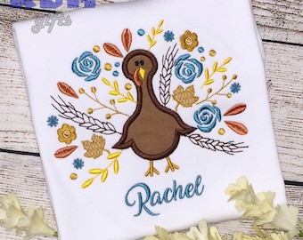 May include: White t-shirt with an embroidered brown turkey surrounded by fall foliage, including wheat, leaves, and flowers in shades of orange, yellow, blue, and gold. The name "Rachel" is embroidered in blue below the turkey.
