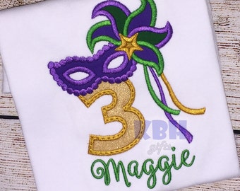 May include: A white t-shirt with a gold glitter number 3, a purple and green Mardi Gras mask, and a green and purple flower. The name "Maggie" is embroidered in green below the number.