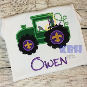 May include: Green tractor with purple wheels and gold fleur-de-lis designs. The tractor is decorated with green, yellow, and purple Mardi Gras beads. The name Owen is embroidered in purple below the tractor.