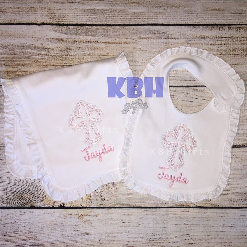 Puede incluir: Un babero y una toallita para eructos de beb&eacute;, ambos blancos con bordes con volantes. Ambos art&iacute;culos presentan una cruz bordada en rosa con pedrer&iacute;a y el nombre "Jayda" en rosa.