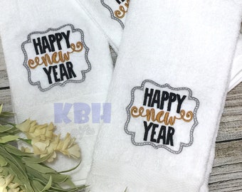 May include: Two white kitchen towels with the text "Happy New Year" embroidered in black and gold lettering within a gray frame.
