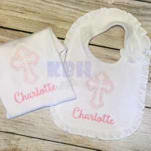Puede incluir: Babero y body de beb&eacute; blancos con una cruz bordada en rosa y el nombre Charlotte.
