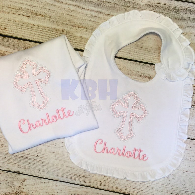 Puede incluir: Babero y body de beb&eacute; blancos con una cruz bordada en rosa y el nombre Charlotte.