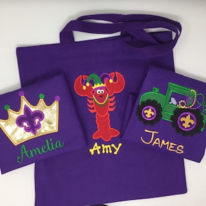 May include: Three purple tote bags with embroidered designs. The first bag has a gold crown with a fleur-de-lis and the name Amelia. The second bag has a red lobster wearing a jester hat and the name Amy. The third bag has a green tractor with fleur-de-lis and the name James.