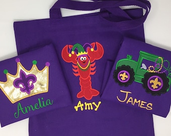 May include: Three purple tote bags with embroidered designs. The first bag has a gold crown with a fleur-de-lis and the name Amelia. The second bag has a red lobster wearing a jester hat and the name Amy. The third bag has a green tractor with fleur-de-lis and the name James.