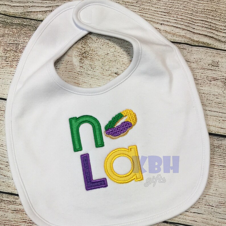 May include: White baby bib with the words "NoLa" embroidered in green, purple, and yellow. A purple, green, and yellow beignet is embroidered above the letters.