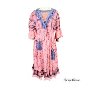 May include: A pink floral print dress with a plaid bodice and blue accents. The dress features a V-neck with blue fringe, bell sleeves, and pockets. The dress has a ruffled hem and is a unique, vintage-inspired design.