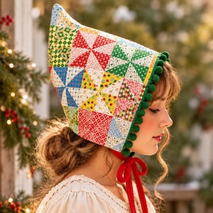 May include: A pointed bonnet featuring a colorful patchwork quilt pattern with red, green, blue, and yellow floral and geometric prints. The edge is trimmed with small green pom-poms, and it secures under the chin with a bright red velvet ribbon.