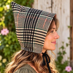 May include: A quilted bonnet featuring a black, white, and green plaid pattern with thin red accent lines. The headwear has a pointed top silhouette and is secured under the chin with black fabric ties that are tied in a simple bow.