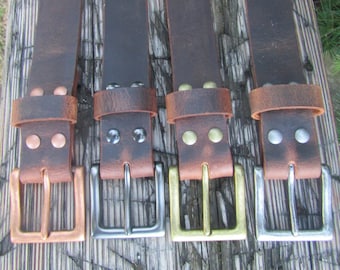 May include: Four dark brown leather belts with matching metal buckles and rivets. Each belt features a distinct metallic finish on the buckle and hardware, including copper, gunmetal, antique brass, and silver tones, arranged in a row.