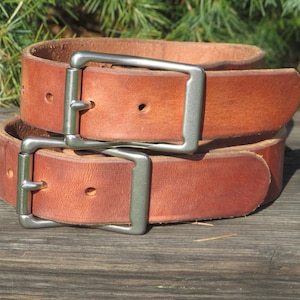 May include: Two tan leather belts coiled together, each featuring a rectangular brushed silver metal buckle with a single prong. The belts have visible stitching holes and a smooth, natural grain finish on the surface of the leather.