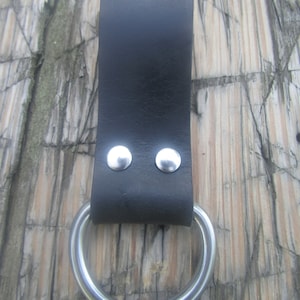 May include: A black leather belt loop with a large, silver-colored metal ring attached. The leather is secured with two silver rivets. The background is a weathered wooden surface.