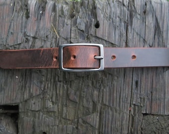May include: A brown leather belt with a silver buckle. The belt is lying on a piece of weathered wood.
