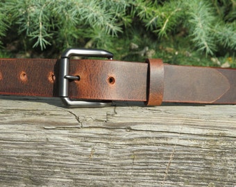 May include: A brown leather belt with a silver-colored buckle. The belt has a distressed appearance with visible grain and a slightly worn look. The background features green foliage and a wooden surface.