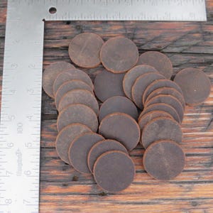 May include: A collection of dark brown, circular leather pieces arranged on a wooden surface. A metal ruler with inch measurements is in the frame, providing scale. The leather circles appear to be approximately 1 inch in diameter.