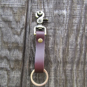 May include: A brown leather key chain with a brass swivel clasp and a brass ring. The key chain is hanging on a wooden surface.