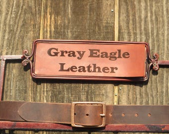 May include: A brown leather belt with a copper buckle and a sign that reads "Gray Eagle Leather." The sign is framed in a decorative copper metal. The background is a wooden surface.