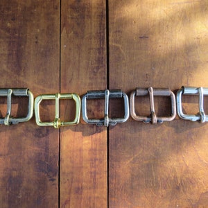 May include: Six metal belt buckles in different finishes: silver, gold, black, copper, and two shades of brass. The buckles are arranged in a row on a wooden surface.