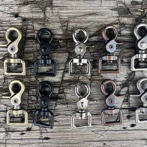 May include: Ten swivel snap hooks in various finishes, including antique brass, black, nickel, and copper. The hooks are arranged in two rows of five on a wooden surface.
