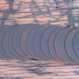 May include: A stack of 18 round, dark brown leather coasters. The coasters are arranged in a semi-circle shape on a wooden surface.