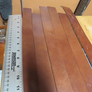 May include: Several long, rectangular strips of smooth, medium-brown leather are arranged side-by-side. A metal Johnson ruler is placed vertically along the left edge, showing measurements in inches up to 11 inches and centimeters up to 28 cm.