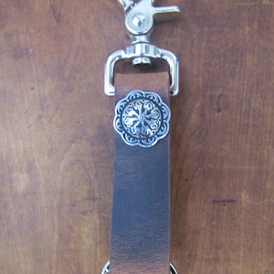 May include: A brown leather keychain with a silver-colored clasp and key ring. The leather strap features a decorative silver-colored embellishment. The keychain is designed for holding keys.