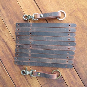 May include: Ten brown leather keychains with a silver swivel clasp and a key ring. The keychains are stacked on top of each other. One keychain has the initials "JKO" engraved on it.