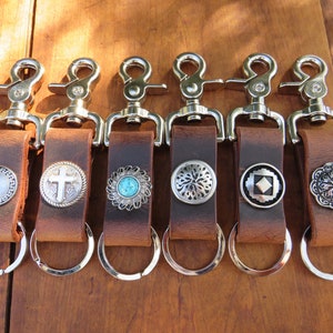 May include: Six brown leather keychains with silver-tone hardware and decorative metal accents. Each keychain features a different design, including a cross, turquoise stone, and geometric patterns. The keychains are displayed on a wooden surface.