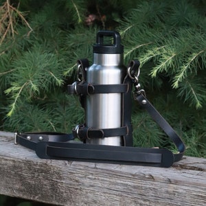 May include: A stainless steel water bottle with a black leather carrying strap. The strap has a buckle and is adjustable. The bottle is sitting on a wooden surface.