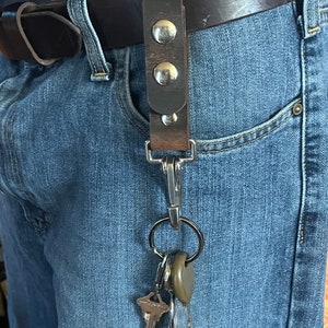May include: A brown leather key chain with silver metal accents and a silver metal clip attached to a belt loop. The key chain has a silver metal ring with a set of keys attached.