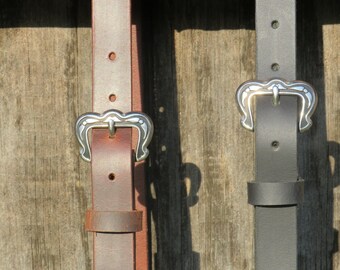 May include: Two leather belts with silver buckles. One belt is brown, the other is gray. The buckles have a decorative, curved design. The belts are hanging against a weathered wooden background.