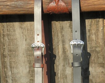 May include: Two leather belts, one brown and one black, hang against a weathered wooden background. Each belt features a silver-colored buckle with an ornate design. The belts are suspended from a rustic metal fixture, suggesting a Western or equestrian theme.