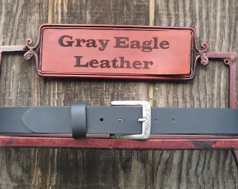 May include: A black leather belt with a silver buckle. The belt is displayed on a red metal stand against a wooden background. A sign above the belt reads "Gray Eagle Leather."
