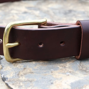 May include: A brown leather belt with a brass buckle. The belt has multiple holes for adjusting the fit.