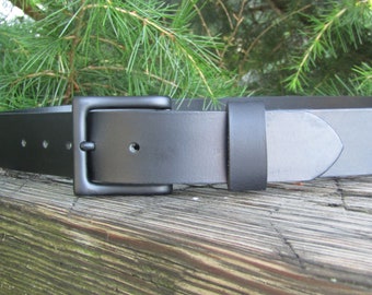 May include: A black leather belt with a black metal buckle. The belt is on a wooden surface.