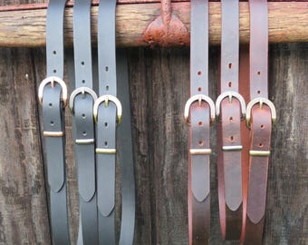 May include: A collection of leather belts in shades of black and brown, each with a brass buckle. The belts are hanging from a rustic, wooden beam with a metal hook, set against a weathered wooden background.