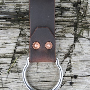 May include: A brown leather belt loop with a silver metal ring. The loop has two copper rivets.