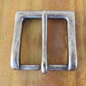 May include: A rectangular, antique-style belt buckle with a tarnished, metallic finish. The buckle has a square frame with a central bar for the belt strap. The metal has a weathered appearance, suggesting age or vintage style.