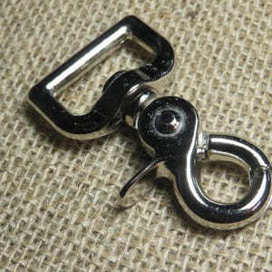 May include: A shiny silver swivel snap hook with a rectangular loop and a spring-loaded clasp. The metal hardware is designed for attaching to straps or other accessories. The hook is shown against a textured, tan background.