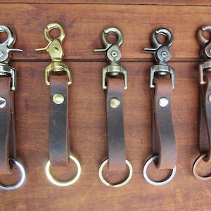May include: Five leather keychains with different metal hardware. Each keychain has a brown leather strap attached to a metal clasp and a metal ring. The clasps are silver, gold, and bronze-toned.