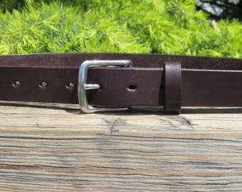 May include: A close-up of a brown leather belt with a silver-colored buckle. The belt has several holes for adjustment and a loop to secure the end. The background features green foliage and a wooden surface.