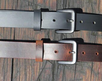 May include: Two leather belts with silver-colored buckles. One belt is black, and the other is brown. The belts are lying on a wooden surface. The buckles are rectangular and have a single prong.